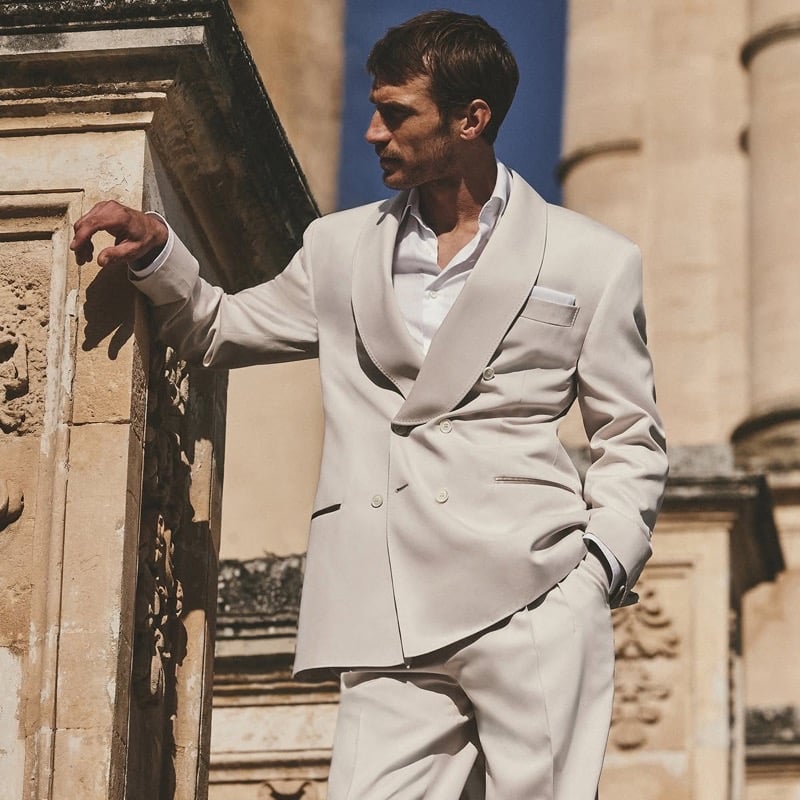 Clément Chabernaud leans against carved stonework in a stone double-breasted shawl-collar suit worn over an open white shirt, set against the bright blue Sicilian sky