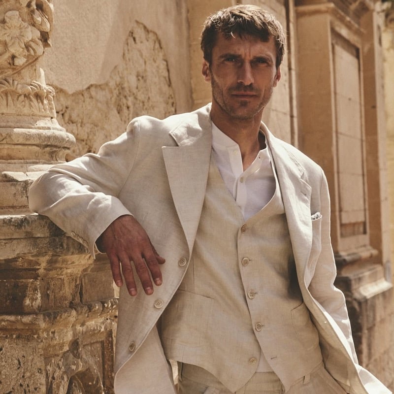 Clément Chabernaud leans against carved limestone in a stone linen three-piece suit with peak lapels over a white granddad-collar henley