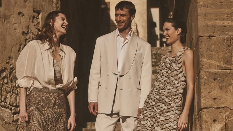 Clément Chabernaud in a stone double-breasted linen suit walks down a Modica alleyway between two female models in scaled metallic dresses, all three laughing together