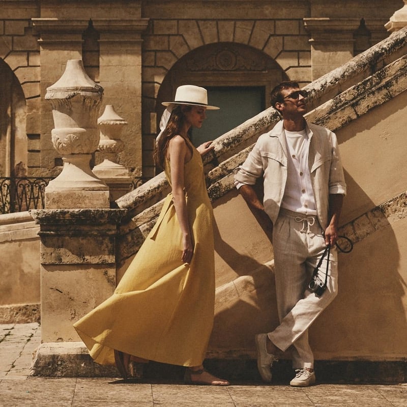 Clément Chabernaud in a stone linen utility shirt-jacket and drawstring trousers leans against a sunlit Modica staircase beside a female model