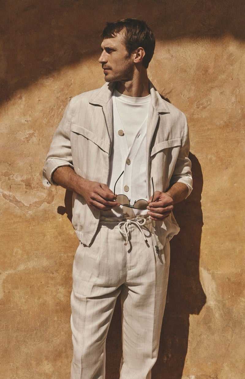 Clément Chabernaud stands against a sun-baked ochre wall in a stone striped linen utility jacket, cardigan, and drawstring trousers over a fine T-shirt, holding sunglasses