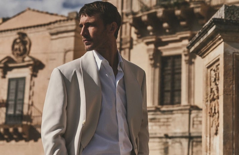 Clément Chabernaud stands on a Modica rooftop in a stone soft-shouldered blazer worn open over a white poplin shirt, baroque architecture behind him