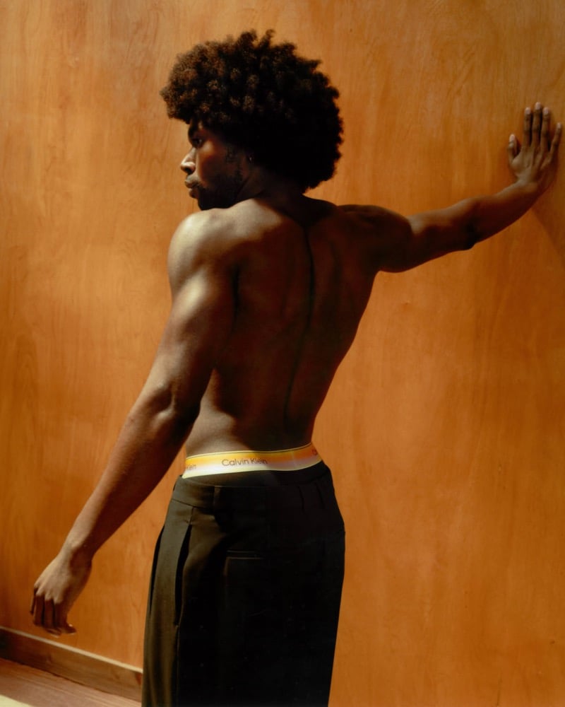 Deon Hinton seen from behind against a warm wood-paneled wall, pressing one hand to the wall in black Calvin Klein trousers with a gradient Pride waistband visible at the back