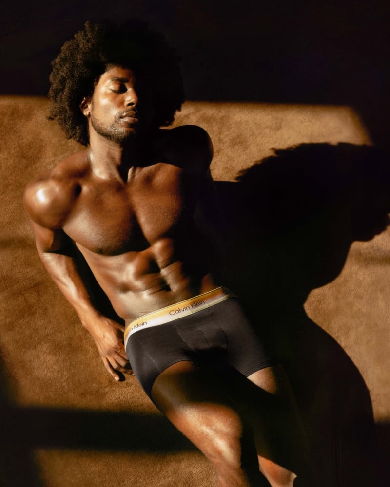 Overhead self-portrait of Deon Hinton reclining on warm brown carpet in black Calvin Klein trunks with a gradient yellow-and-white waistband, eyes closed in a wash of low amber light