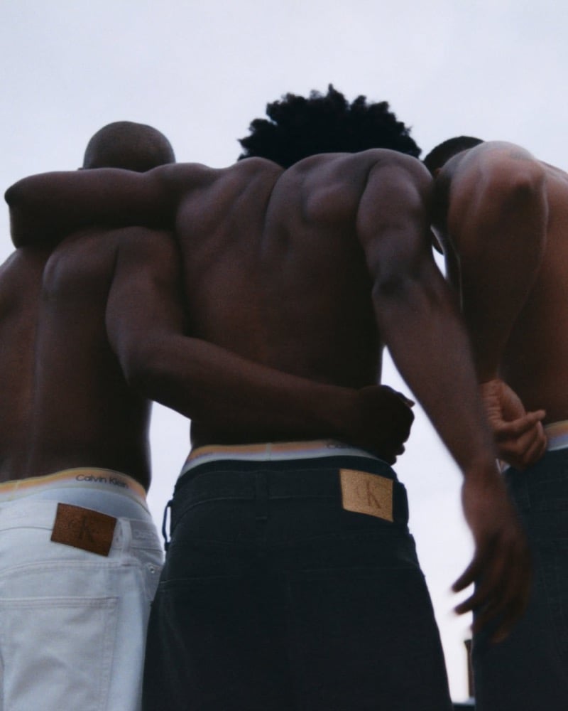Low-angle shot of three shirtless men photographed from behind with arms wrapped around each other's shoulders against an overcast sky, wearing Calvin Klein Pride underwear under white and black CK monogram-patched jeans
