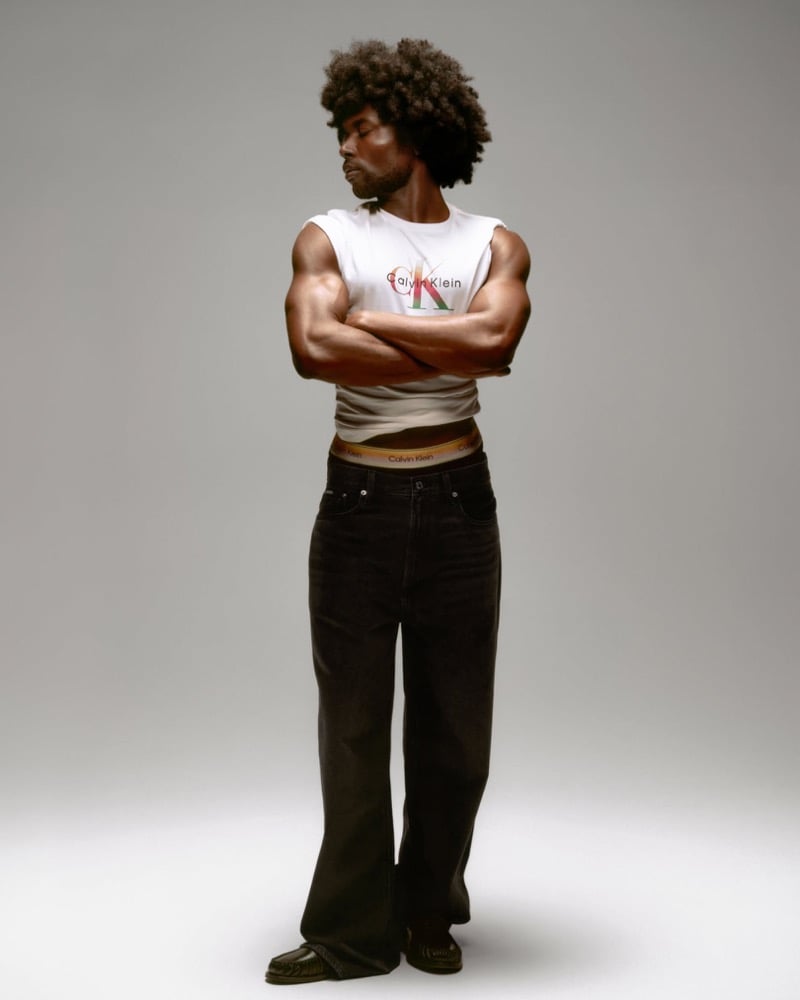 Deon Hinton stands with arms crossed in a fitted white Calvin Klein logo t-shirt printed with a rainbow CK monogram, layered over a gradient Pride waistband peeking above black wide-leg jeans