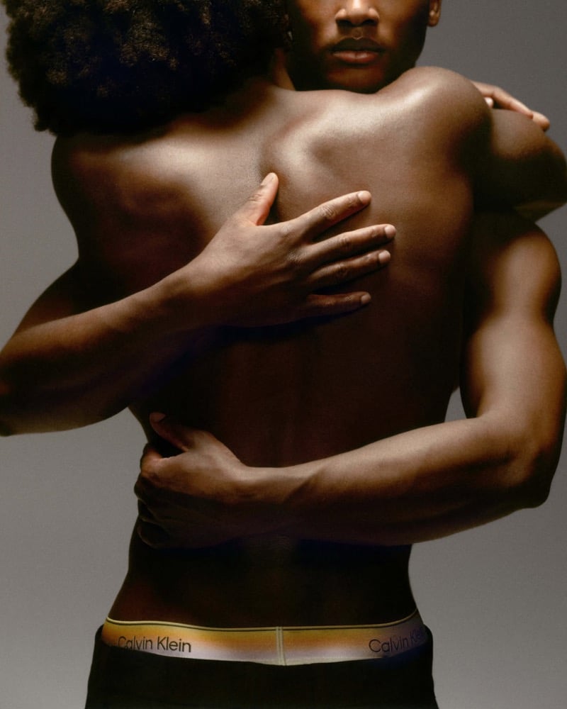 Close cropped shot of two men in a tight embrace from the shoulders down, both wearing black Calvin Klein Pride trunks with rainbow gradient waistbands against a warm gray studio backdrop