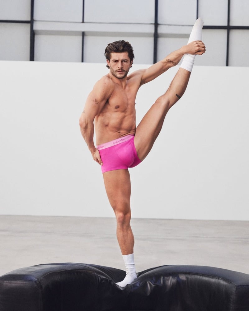 Sam Salter balances on a black leather bench in hot pink Calvin Klein Pride trunks and white tube socks, extending one leg vertically toward the rafters of a sunlit studio