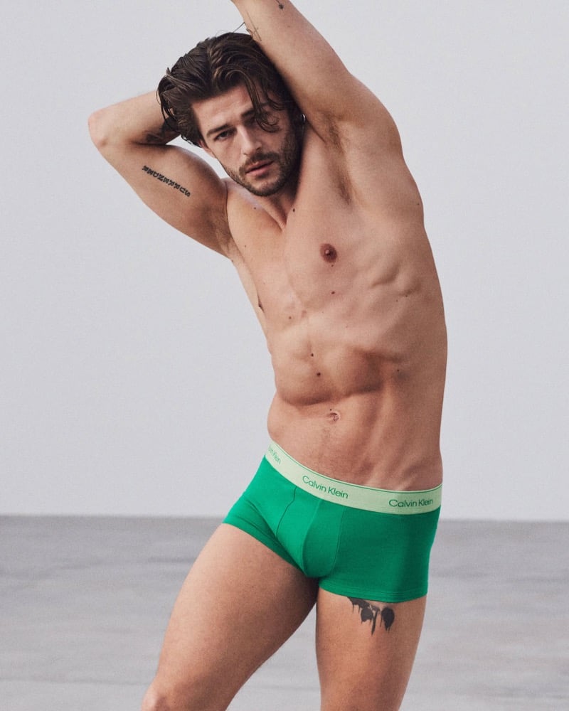 Sam Salter raises both arms above his head with damp hair falling forward, shirtless in green Calvin Klein Pride trunks with a pale gradient waistband, against a soft gray studio backdrop