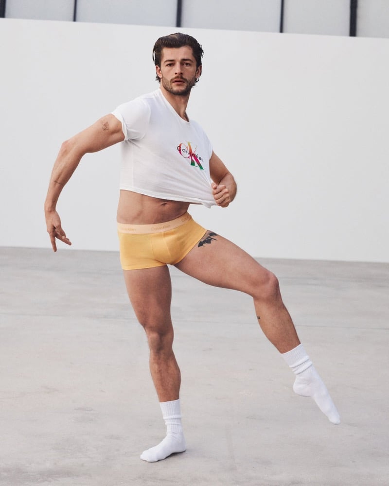 Sam Salter mid-stride on a concrete studio floor in white tube socks and yellow Calvin Klein Pride trunks with a pale waistband, lifting the hem of a white Calvin Klein logo tee printed with a rainbow CK monogram