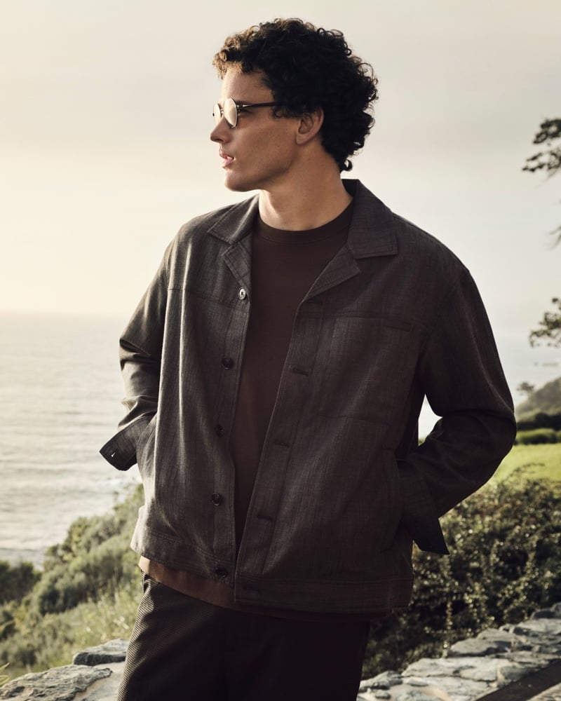 Simon Nessman wears an oversized brown overshirt over a brown crewneck with dark trousers and sunglasses, photographed against a cliffside ocean view