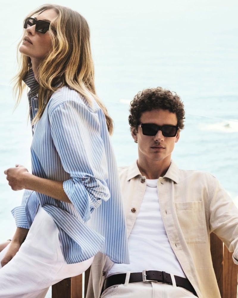 Simon Nessman wears a lightweight beige overshirt over a tee with a leather belt and dark sunglasses, beside a partner in a blue-striped shirt by the ocean