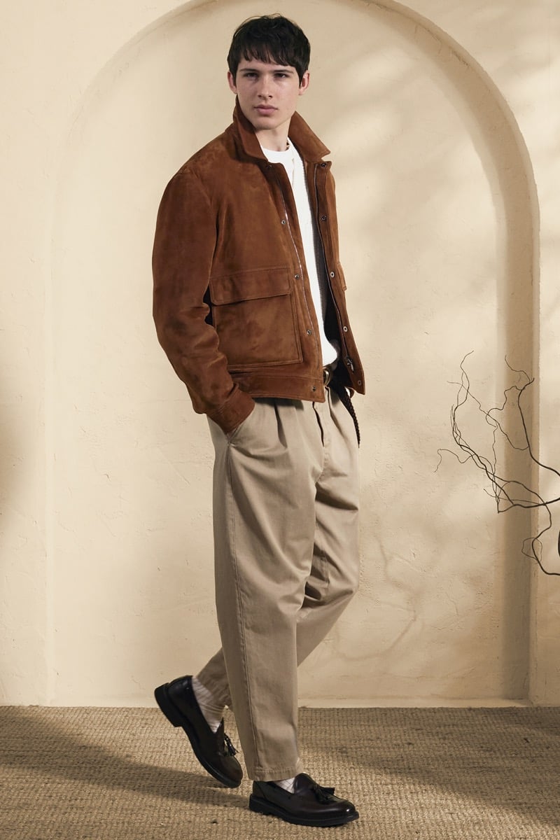 Model Mateo Ruiz in a cognac suede blouson with patch pockets over a cream ribbed crewneck, paired with khaki pleated trousers and dark brown tassel loafers