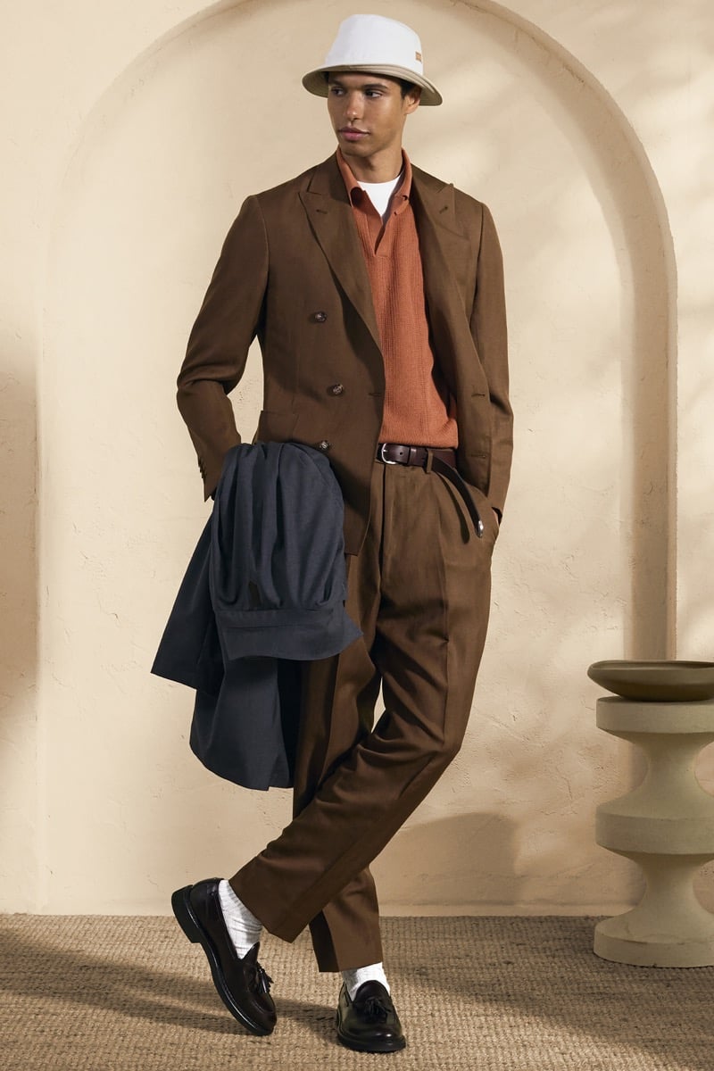 Model Gilbert van Damme in a cream and tan panama hat with a brown band wears a chocolate brown double-breasted suit over a rust orange knit polo