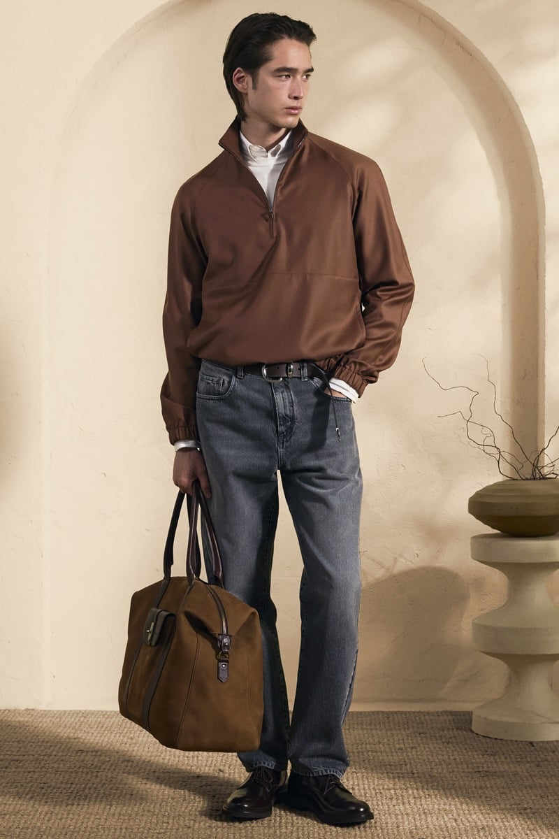 Model Peter Meyer in a chocolate brown half-zip pullover over a white shirt, paired with gray washed denim jeans and a brown suede weekender bag