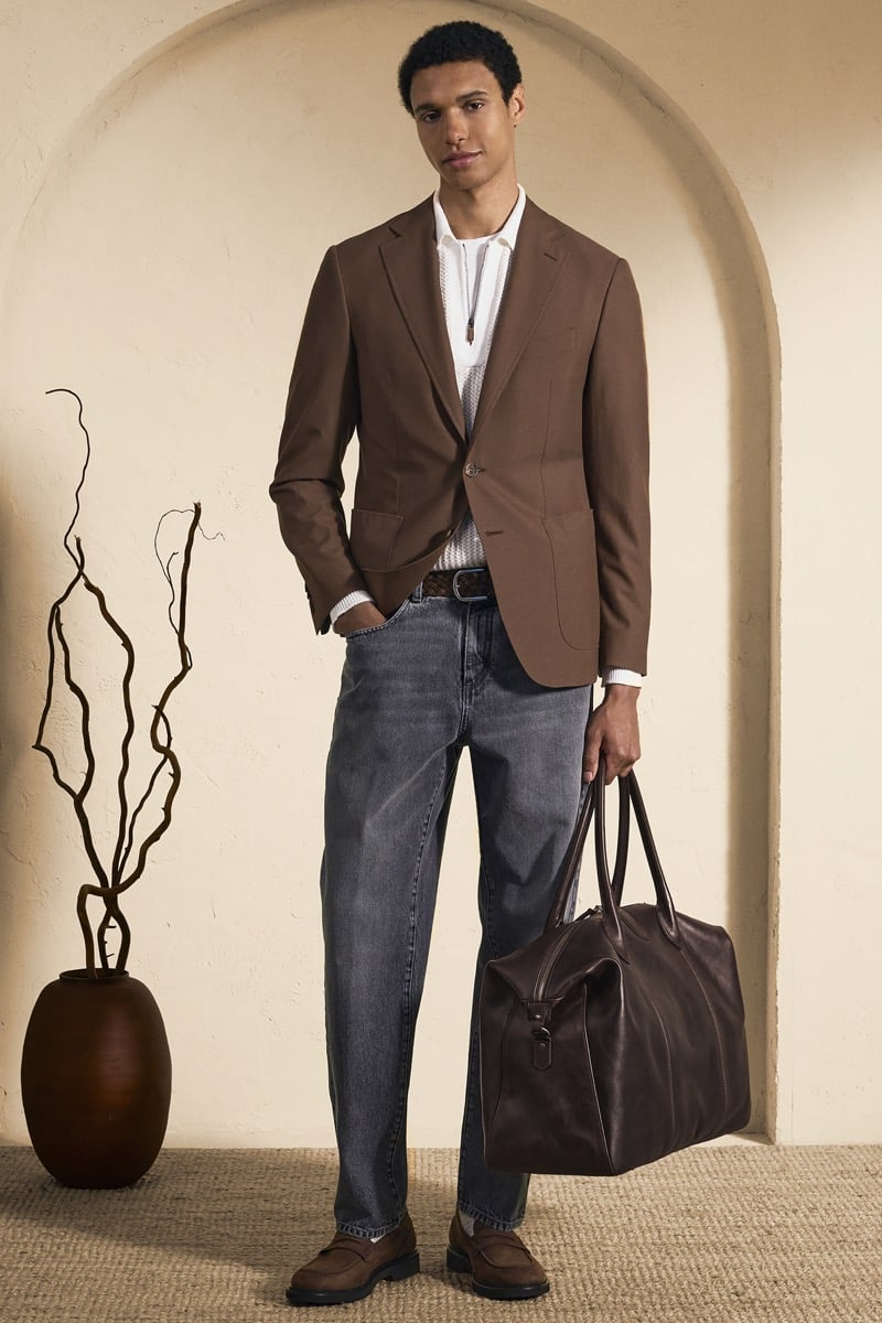 Model Gilbert van Damme in an unstructured chocolate brown blazer over a white openwork knit polo and woven braided belt, paired with gray washed jeans and a dark brown leather weekender