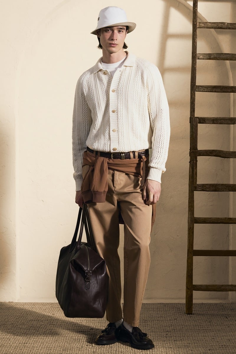 Model Peter Meyer in a white bucket hat wears a cream openwork knit cardigan over a tan jacket tied at the waist and pleated tobacco trousers, carrying a dark brown leather holdall