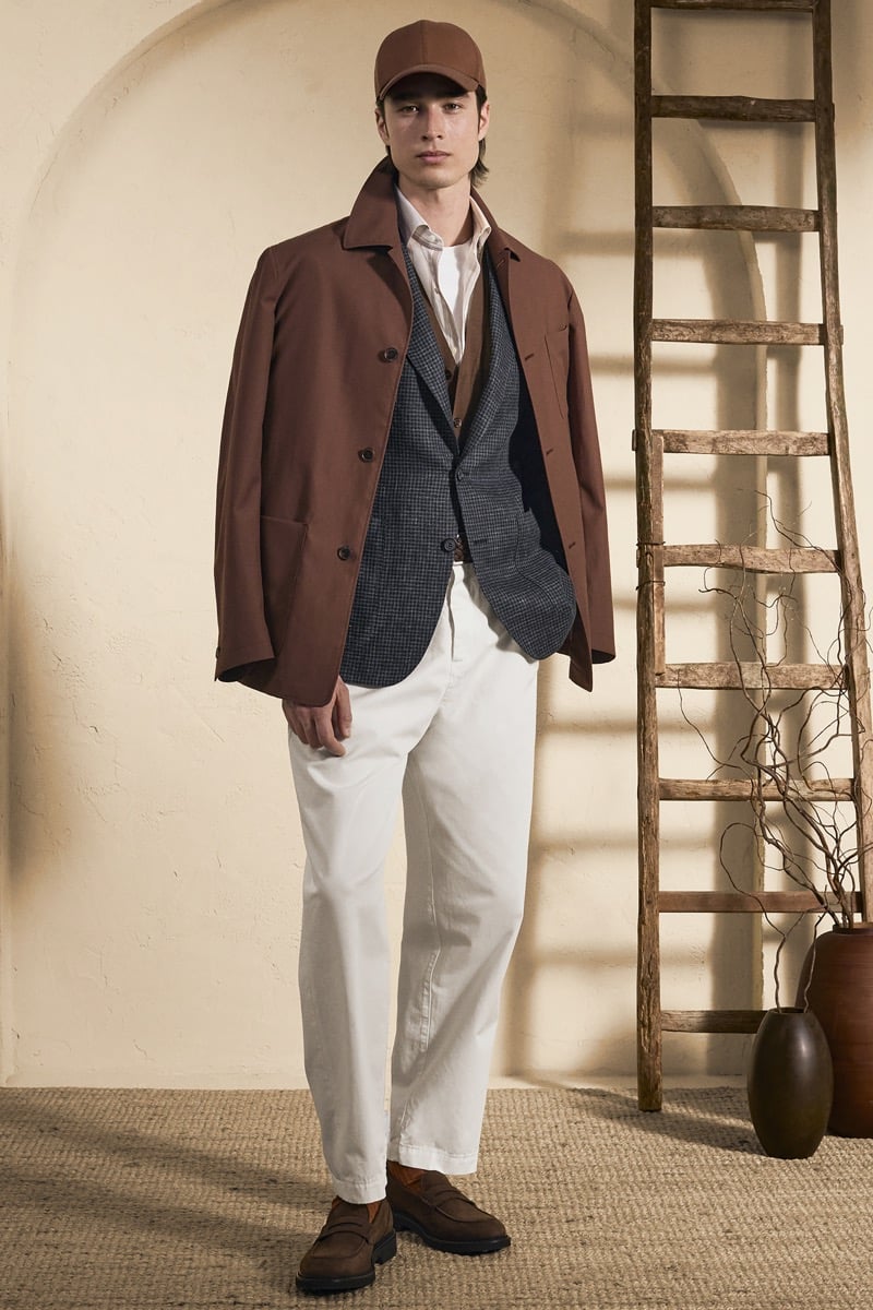 Model Peter Meyer in a brown baseball cap wears an unstructured rust brown coat layered over a navy houndstooth blazer and white shirt, paired with cream wide-leg trousers and brown suede loafers