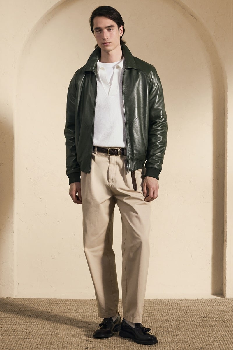 Model Peter Meyer in a dark green leather harrington jacket over a white openwork knit polo, paired with stone pleated trousers, a braided brown belt, and black tassel loafers
