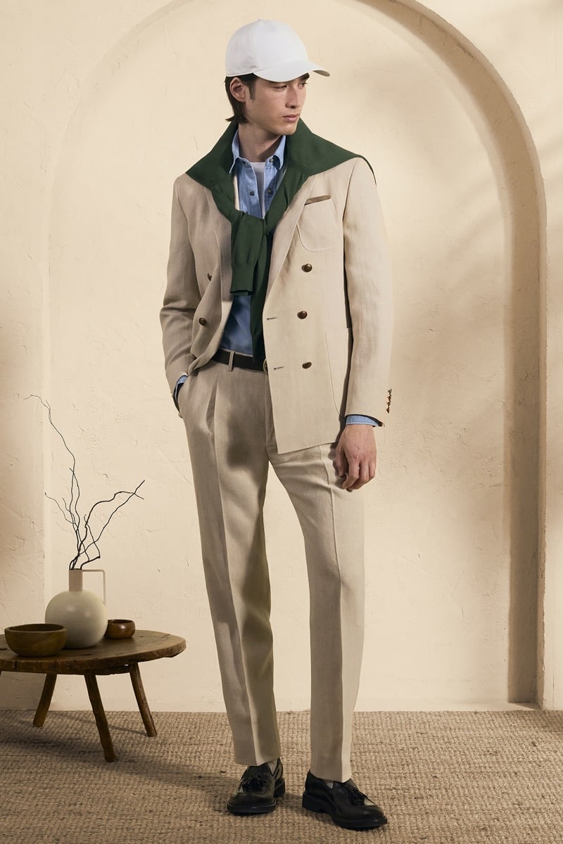 Model Peter Meyer in a white baseball cap wears a cream linen double-breasted suit over a light blue chambray shirt with a green knit sweater draped across the shoulders