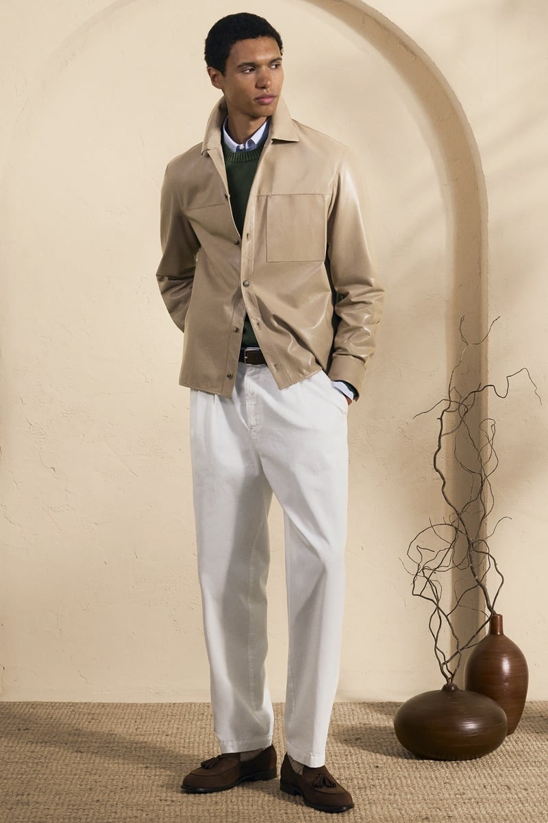 Model Gilbert van Damme in a sand leather shirt jacket over a forest green sweater and pale blue button-down, paired with white trousers and brown suede loafers