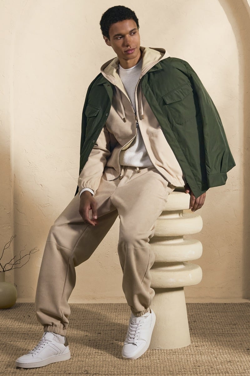 Model Gilbert van Damme in a forest green jacket over a sand zip hoodie and white ribbed knit, paired with matching sand sweatpants and white high-top sneakers