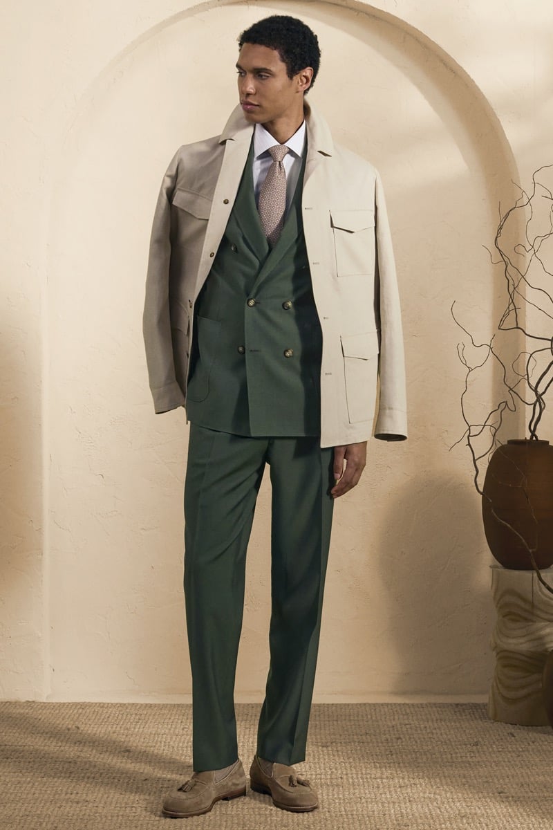Model Gilbert van Damme in a beige utility chore jacket over a forest green double-breasted suit, white shirt, and printed silk tie, paired with sand suede tassel loafers