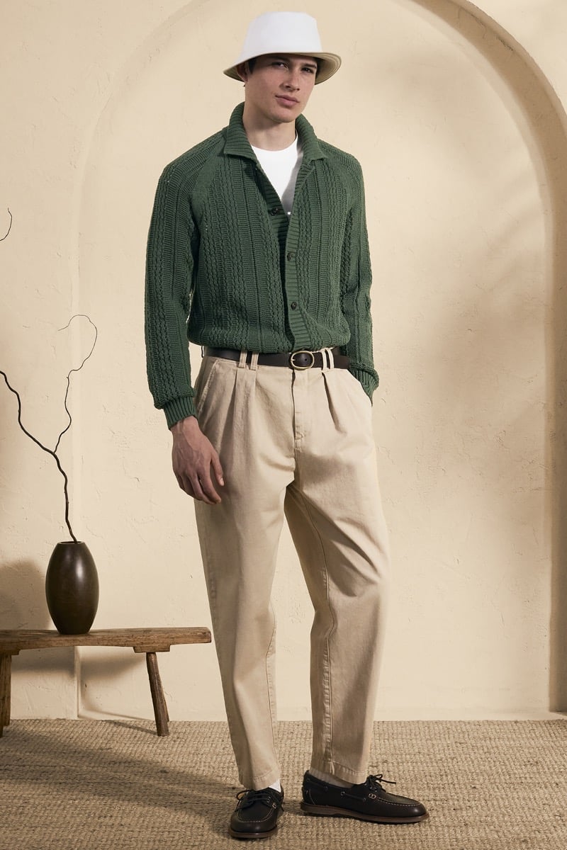 Model Mateo Ruiz in a white bucket hat wears a forest green openwork cable-knit shirt cardigan over a white tee, paired with stone pleated trousers and navy boat shoes