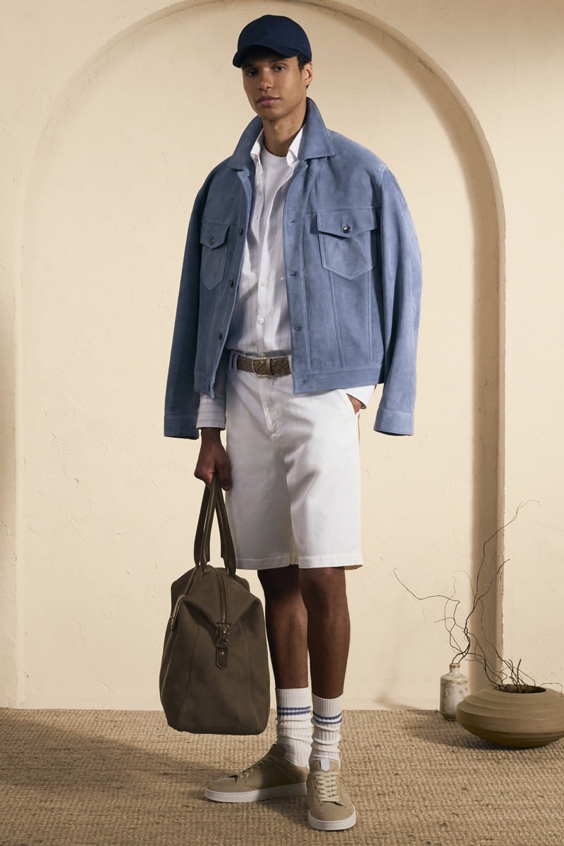 Model Gilbert van Damme in a navy baseball cap wears a sky blue suede trucker jacket over a white shirt and braided belt, paired with white denim shorts and tan suede sneakers