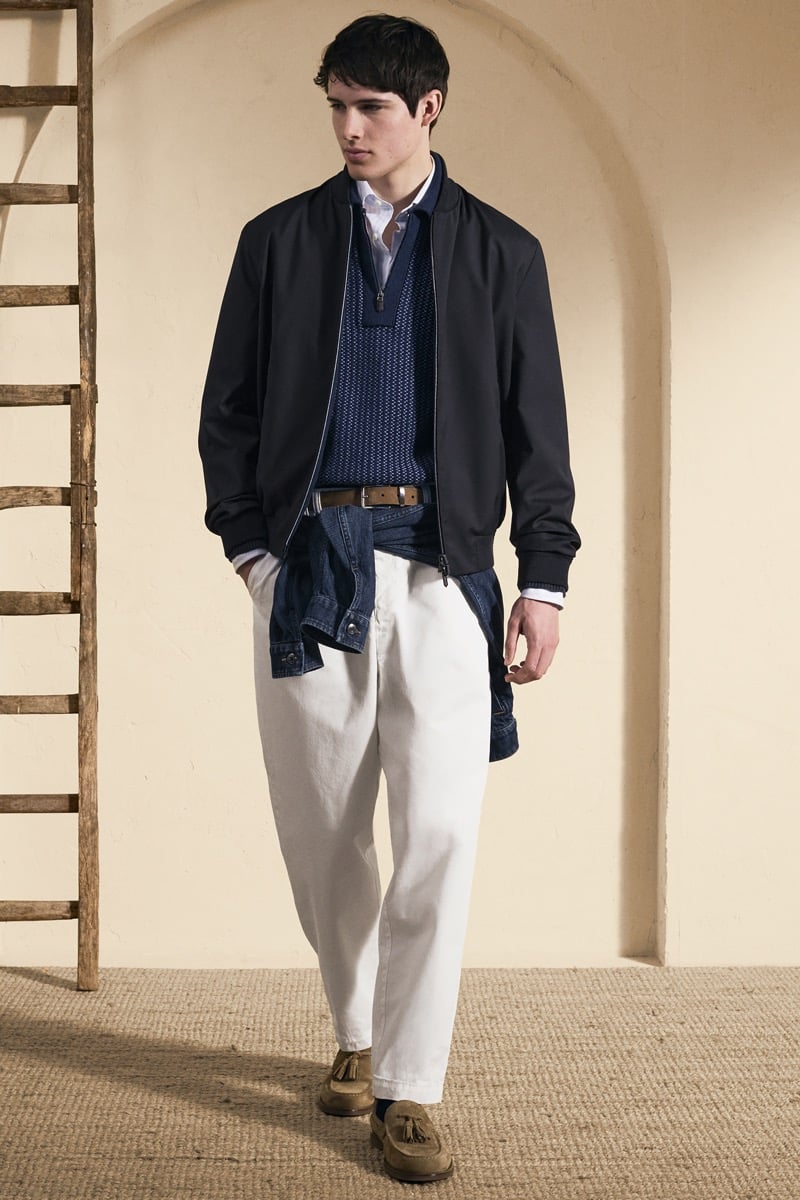 Model Mateo Ruiz in a navy bomber jacket over a navy chevron-knit half-zip sweater and white shirt, with a denim jacket tied at the waist over white pleated trousers and tan suede tassel loafers
