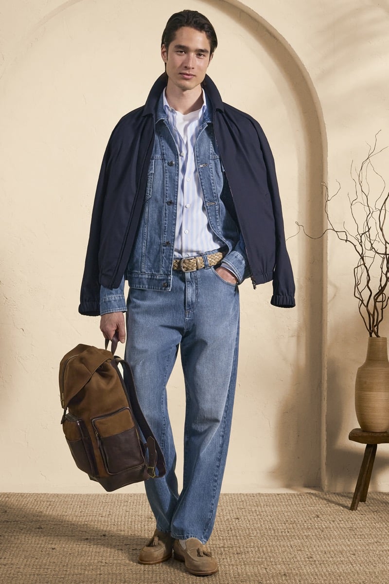 Model Peter Meyer in a navy bomber jacket layered over a denim chore jacket and blue striped shirt, paired with light wash jeans, a woven belt, and a brown suede backpack