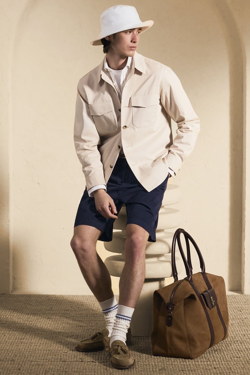 Model Peter Meyer in a white bucket hat wears a cream cotton chore jacket over a white tee, paired with navy chino shorts, striped athletic socks, and tan suede boat shoes with a brown suede holdall