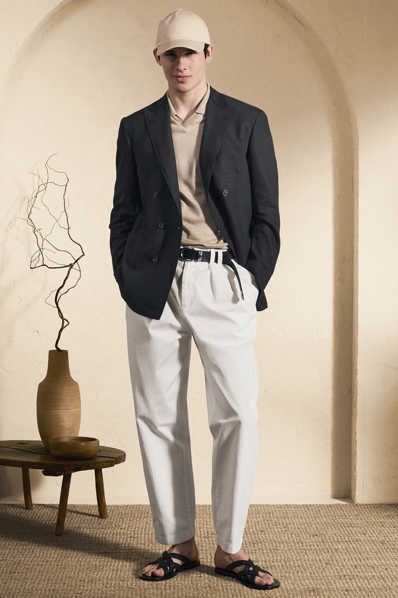 Model Mateo Ruiz in a cream baseball cap wears a black double-breasted linen blazer over a sand knit polo, paired with white pleated trousers, a striped canvas belt, and black leather sandals