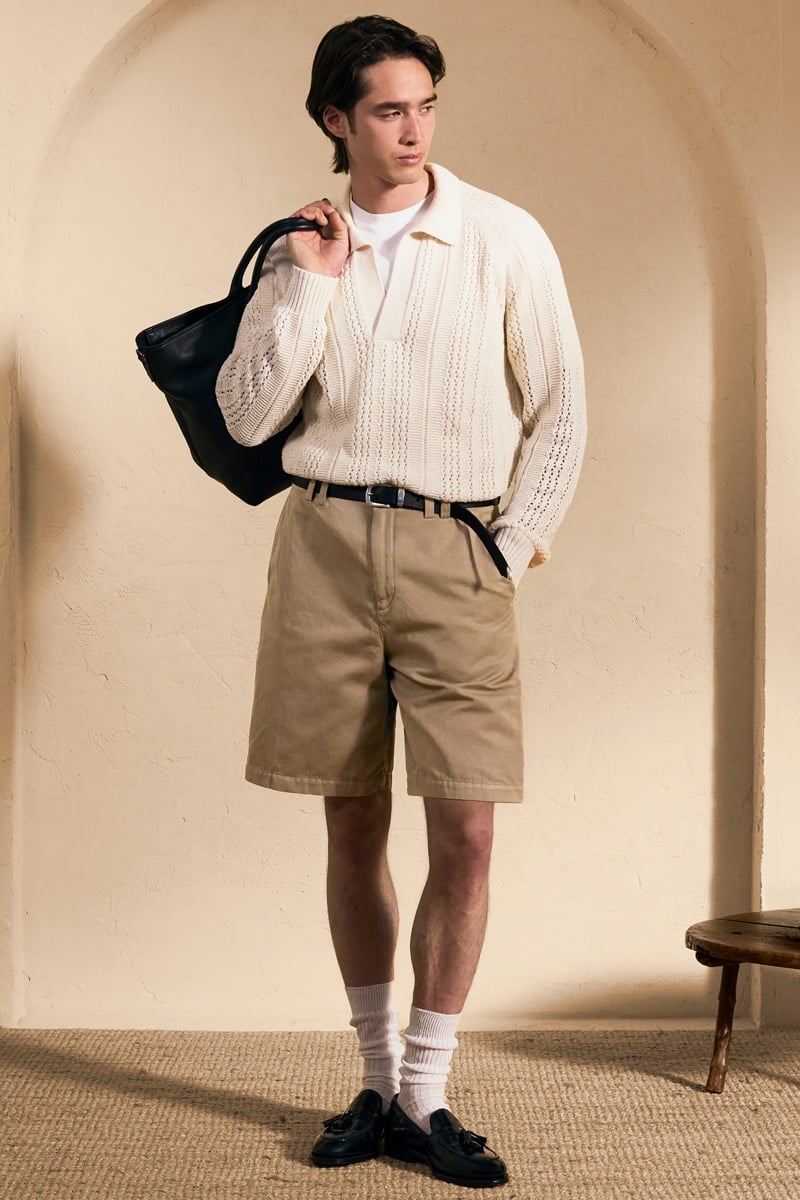 Model Peter Meyer in a cream openwork knit polo with a white tee underneath, paired with khaki pleated shorts, ribbed white socks, and black tassel loafers, holding a black leather tote