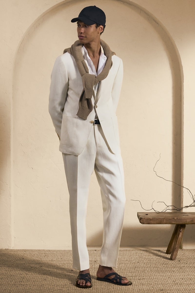Model Gilbert van Damme in a navy baseball cap wears an ivory linen suit over a white shirt with a taupe knit tied around the shoulders, finished with black leather sandals