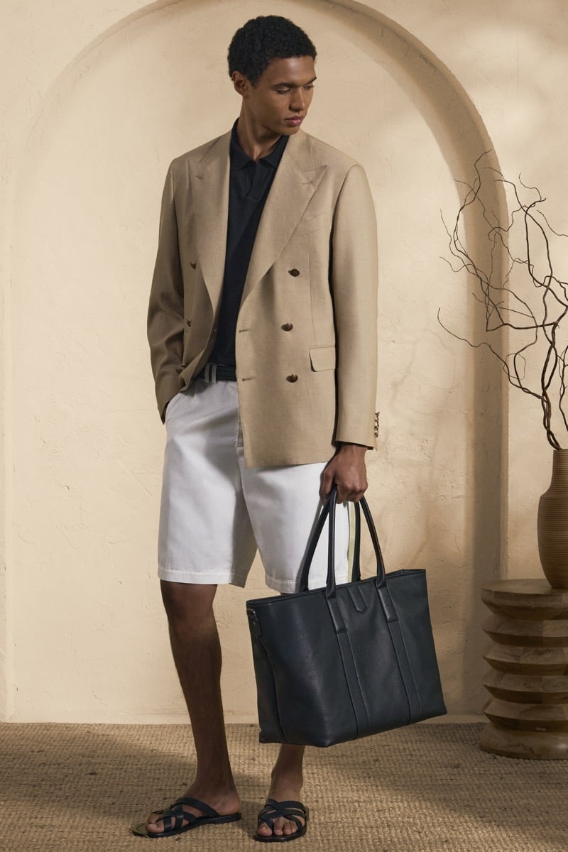 Model Gilbert van Damme in a camel double-breasted blazer over a navy polo, paired with white linen shorts, black leather sandals, and a black leather tote