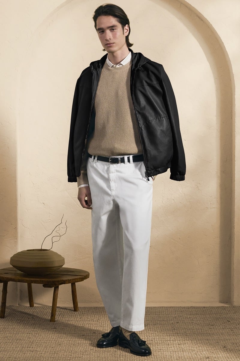 Model Peter Meyer in a black hooded leather jacket over a camel ribbed crewneck and white shirt, paired with white pleated trousers, a canvas belt, and black tassel loafers