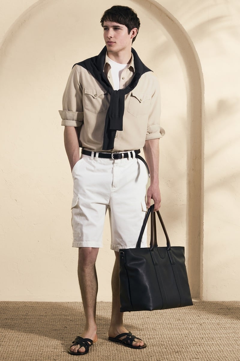 Model Mateo Ruiz in a sand utility shirt with snap pockets layered over a white tee with a black knit tied around the shoulders, paired with white cargo shorts, a belt, and black leather sandals
