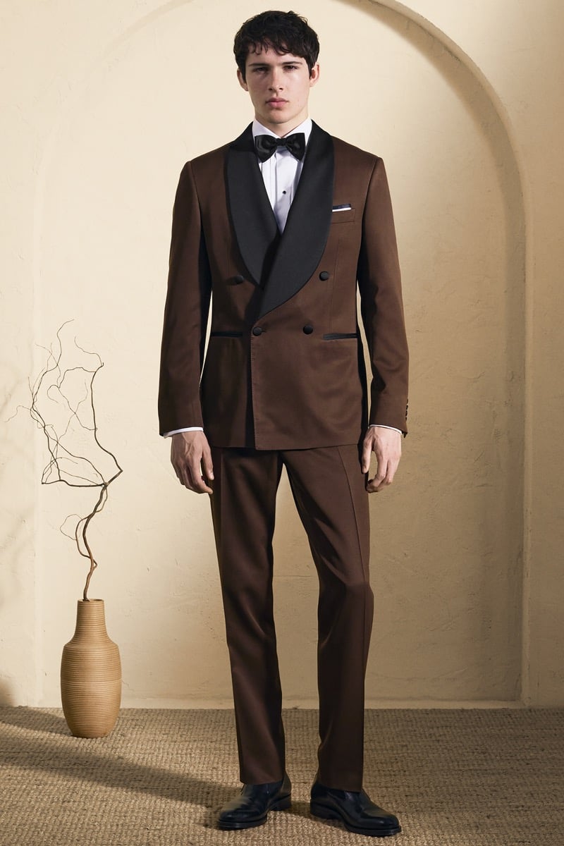 Model Mateo Ruiz in a chocolate brown double-breasted tuxedo with black satin shawl lapels over a white tuxedo shirt and black bow tie, paired with matching brown trousers and black leather loafers