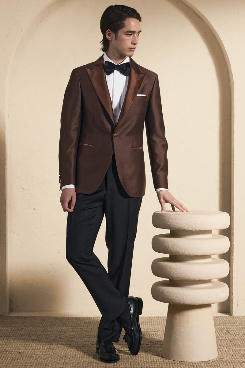 Model Peter Meyer in a copper bronze peak-lapel dinner jacket with tonal satin lapels over a white shirt and navy bow tie, paired with black trousers and navy tassel loafers