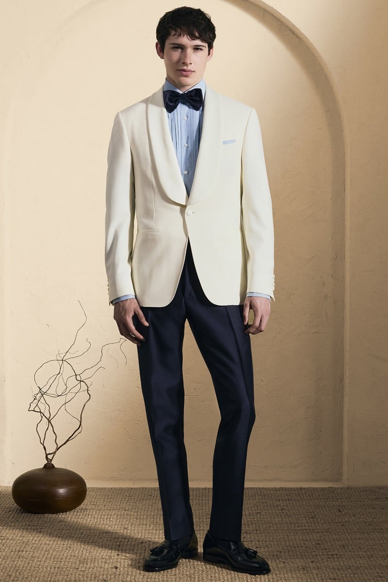 Model Mateo Ruiz in a cream shawl-collar dinner jacket over a pale blue tuxedo shirt with a navy bow tie, paired with navy trousers and black tassel loafers
