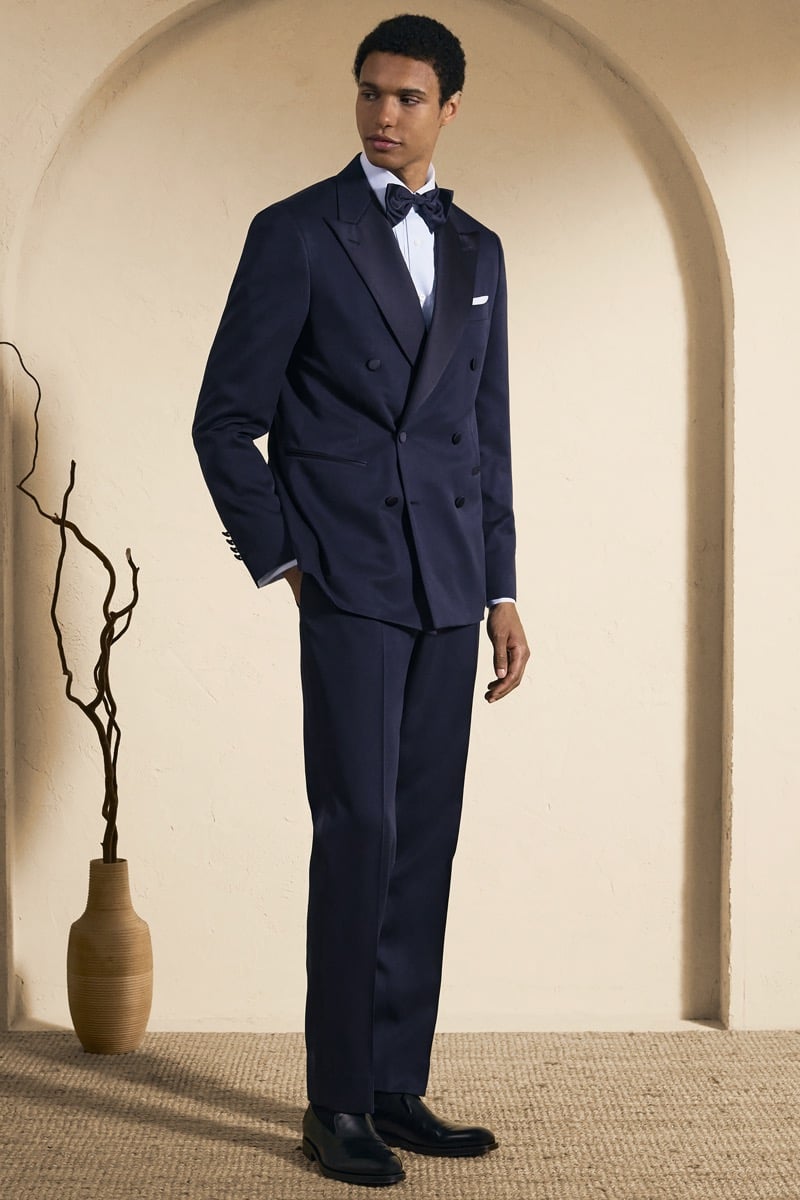 Model Gilbert van Damme in a navy double-breasted peak-lapel tuxedo with satin lapels over a white shirt and navy bow tie, finished with black leather loafers