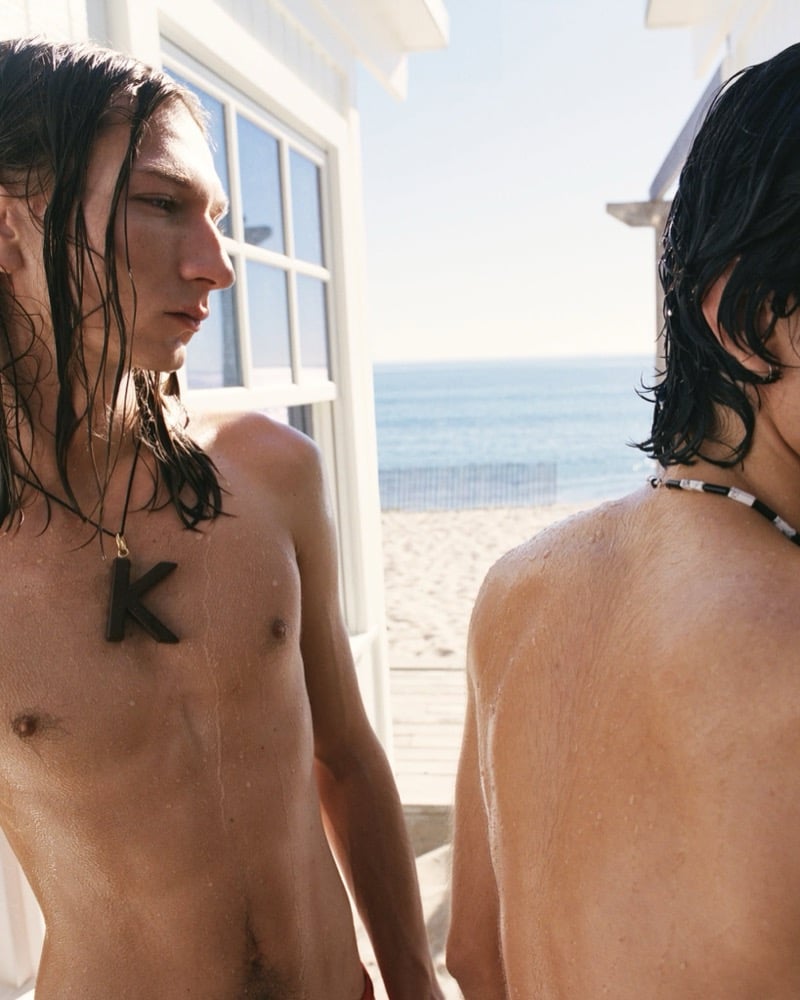Gabriel Tiercelin in profile with a K pendant chain stands beside Samuel Funs wearing a beaded chain, both shirtless against a beach backdrop
