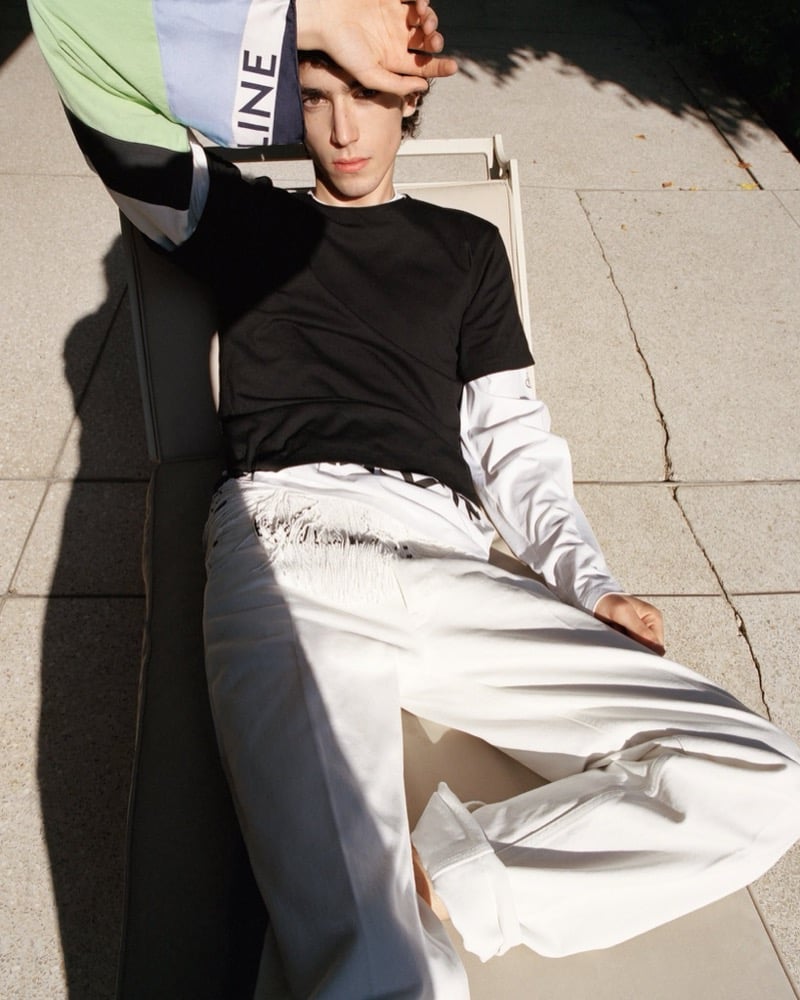 Samuel Funs reclines in a lounge chair in a black tee layered over a Celine colorblock logo shirt with green and blue panels, paired with wide-leg trousers
