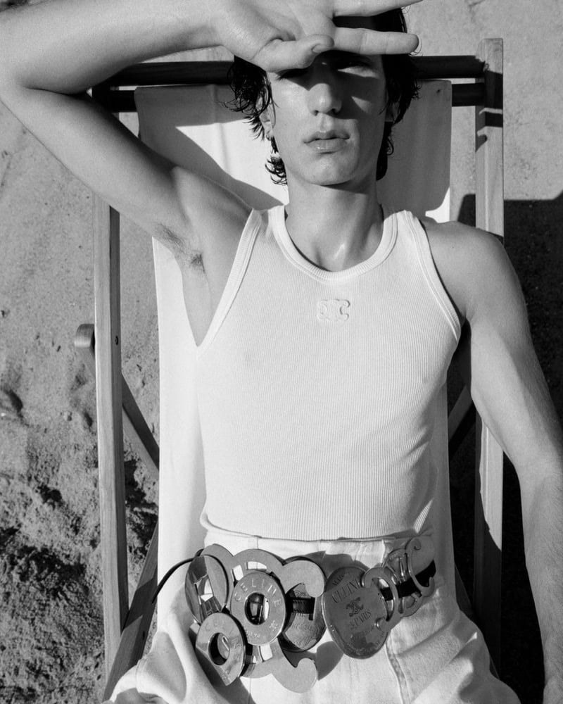 Samuel Funs raises a hand to shade his face, wearing a ribbed Celine logo tank tucked into trousers cinched with an oversized metal link belt