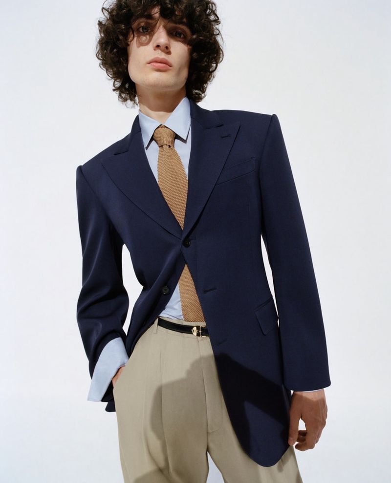 Bas Van Geertruy in a navy single-breasted peak-lapel blazer over a pale blue shirt and tan knit tie, paired with sand pleated trousers and a slim black leather belt with gold buckle