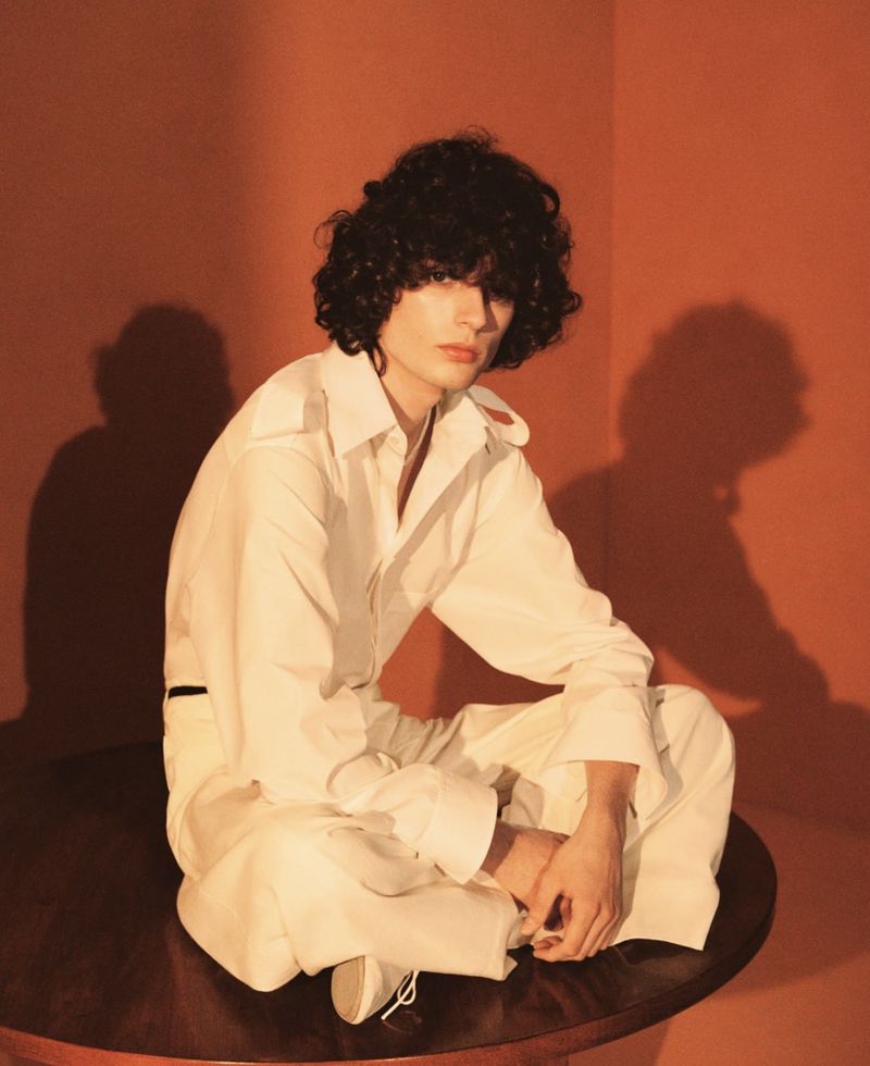 Bas Van Geertruy sits cross-legged on a round dark wooden table in an oversized ivory poplin shirt and matching wide-leg trousers, lit warmly against a terracotta backdrop