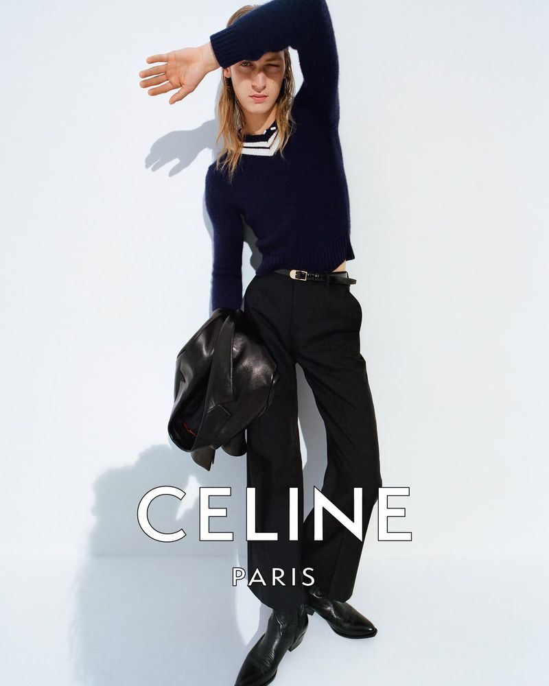 Jesse Rinderknecht in a navy V-neck sweater layered over a striped collared top, paired with dark wide-leg trousers, a slim black belt, black western boots