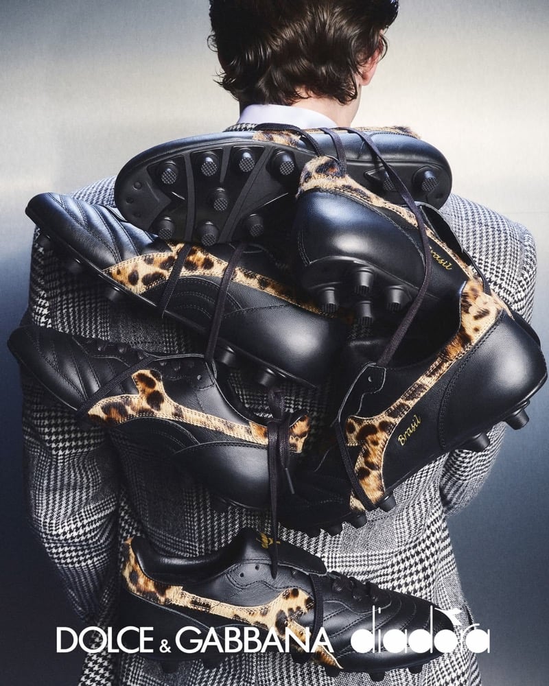 Back view of Max Knott in a Prince of Wales check tailored jacket, holding a stack of Dolce & Gabbana x Diadora Brasil boots and lifestyle sneakers in black leather with leopard pony hair panels, the Dolce & Gabbana and Diadora logos locked up at the foot of the frame