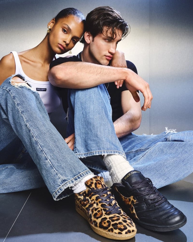 Farah Nieuwburg in a white Dolce & Gabbana ribbed tank top and ripped blue denim leaning into Max Knott in a black tee and faded ripped jeans, both seated on a polished floor with one wearing leopard pony hair Brasil sneakers and the other in leopard and black Brasil leather